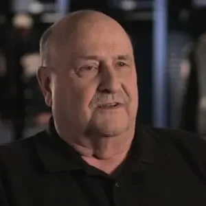 A man in black shirt and beard looking at camera.